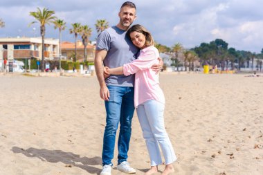 Romantik Kafkasyalı çift sahilde kucaklaşırken birlikte vakit geçiriyorlar. Parlak bir gökyüzünün altında kumla, denizle çevrili bir şekilde birbirlerine sevgiyle gülümserler ve kameraya bakarlar.