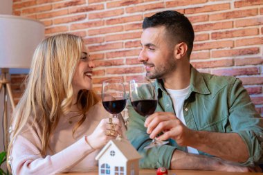 Kırmızı şarapla kadeh kaldıran ve birbirlerine sevgiyle gülümseyen mutlu beyaz çift. Masanın üzerindeki minyatür bir ev ve anahtarlar rahat ve modern bir yerde yeni bir eve taşınmanın neşesini simgeliyor.. 