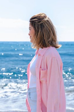 Kumlu bir plajda oturmuş, arkasında okyanus varken kameraya gülümseyen mutlu genç bir kadının dikey portresi. Rahat bir ortam, günlük kıyafetler, doğal ışık ve sakin bir ortam.. 