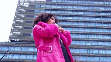 Pembe palto giyen kıvırcık saçlı Afrikalı Amerikalı dansçı şehir ortamında hareketlerini ve enerjilerini ifade ediyor. Modern moda, çağdaş dans ve sanatsal performans şehrin arka planına karşı.