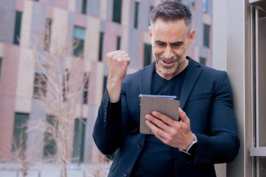 Kafkasyalı olgun bir adam şehir ortamında tabletine bakarken zaferini kutluyor. Yükseltilmiş yumruk ve neşeli ifade dijital ortamda başarı, heyecan ve başarıyı yansıtır 