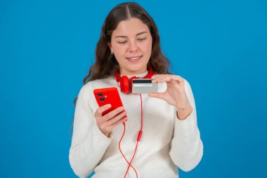 Beyaz kazaklı ve kırmızı kulaklıklı mutlu genç bir kadın kameraya bir kredi kartı gösterirken gülümsüyor ve elinde kırmızı bir akıllı telefon tutuyor, mavi arka planda online alışverişi sembolize ediyor.. 