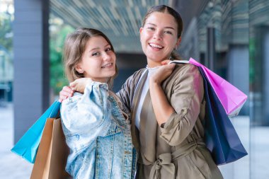 Renkli alışveriş torbaları taşıyan ve alışveriş merkezinde başarılı bir günün ardından gülümseyen iki beyaz kadın. Modern bir binanın önünde duruyorlar, birlikte eğleniyorlar stil ve neşeyle. 