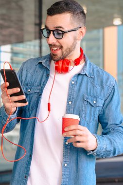 Şehir ortamında akıllı telefonunu kullanırken kırmızı kulaklıklı genç adam kahvenin tadını çıkarıyor. O okurken ya da mesajlaşırken gülümsüyor, modern cam binaların önünde sıradan bir anda duruyor. Dikey