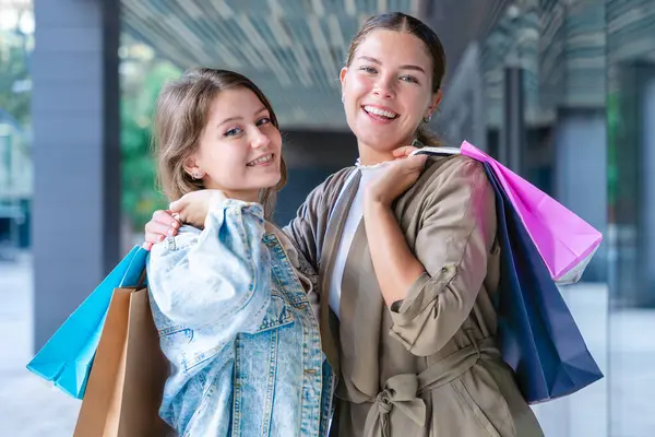 Renkli alışveriş torbaları taşıyan ve alışveriş merkezinde başarılı bir günün ardından gülümseyen iki beyaz kadın. Modern bir binanın önünde duruyorlar, birlikte eğleniyorlar stil ve neşeyle. 