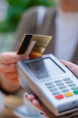 Yakından biri, POS terminalindeki bir kredi kartıyla bağlantısız ödeme yapmış. Sahne arka planında çaydanlık olan bir kafe ortamında geçiyor, modern işlem kolaylığını gösteriyor.