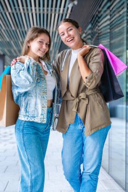 Renkli alışveriş torbaları taşıyan ve alışveriş merkezinde başarılı bir günün ardından gülümseyen iki beyaz kadın. Modern bir binanın dışında duruyorlar, birlikte eğleniyorlar stil ve neşe içinde. Dikey