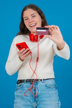 Beyaz kazaklı ve kırmızı kulaklıklı mutlu genç bir kadın kameraya bir kredi kartı gösterirken gülümsüyor ve elinde kırmızı bir akıllı telefon tutuyor, mavi arka planda, online alışverişi sembolize ediyor. Dikey