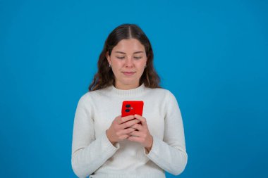 Genç bayan beyaz süveter, kırmızı akıllı telefonuna tarafsız ya da biraz düşünceli bir ifadeyle bakıyor, sağlam bir mavi arkaplanda duruyor, odak ve dijital bağlantıyı temsil ediyor.. 