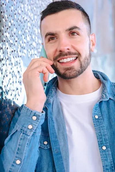 Açık havada akıllı telefondan konuşan yakın plan gülümseyen adam. Bir kot ceket giyiyor ve yansıtıcı bir yüzeye yakın duruyor, doğal gün ışığında gündelik ve modern bir iletişim anının tadını çıkarıyor. Dikey