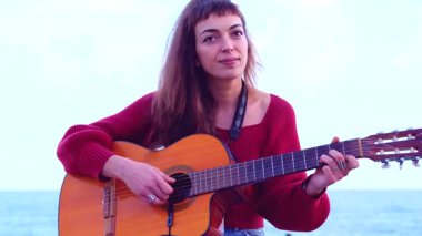 Akustik gitar çalan ve deniz kenarında şarkı söyleyen sanatçı genç bir kadın. Doğayla ve yumuşak doğal ışıkla çevrili sakin bir sahil ortamında duygusal bir performans sergiliyor.. 