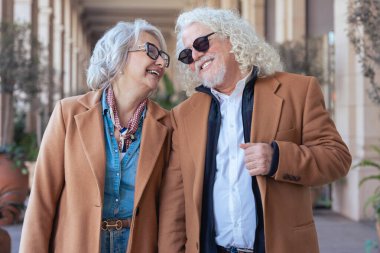Güneş gözlüğü ve şık ceketler takan gülümseyen yaşlı bir çiftin dikey portresi. Şehir ortamında zarafet, mutluluk ve bağlantı göstererek güvenle dışarı çıkarlar.. 
