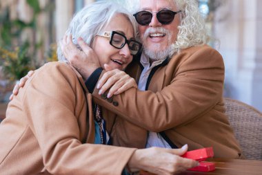 Gözlüklü ve kahverengi paltolu yaşlı bir çift açık hava kafesinde kahvenin tadını çıkarıyor. Sıcak bir anı paylaşıyorlar, gün içinde huzurlu ve rahat bir atmosferde gülümsüyorlar. Dikey
