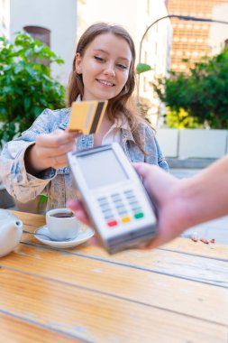 Açık bir kafede temassız teknoloji kullanan beyaz bir kadın kredi kartıyla ödeme yaparken gülümsüyor. Rahat, yaşam tarzı ve modern finansal alışkanlıkları yansıtan sıradan bir kentsel an. Dikey