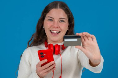 Beyaz kazaklı ve kırmızı kulaklıklı mutlu genç bir kadın kameraya bir kredi kartı gösterirken gülümsüyor ve elinde kırmızı bir akıllı telefon tutuyor, mavi arka planda online alışverişi sembolize ediyor.. 
