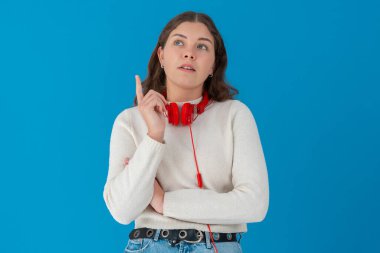 Boynunda kırmızı kulaklık olan beyaz kazaklı genç bir kadın sanki bir şey hatırlıyormuş ya da anlıyormuş gibi parmağını kaldırıyor. Düşünceli, mavi arka planda izole bir şekilde yukarı bakıyor. Boşluğu kopyala. 