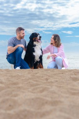 Tatil sırasında kumsalda büyük bir köpekle gülümseyen beyaz bir çiftin portresi. Deniz kenarında rahat ve neşeli bir an, üçünün de parlak bir günde kameraya baktığı. Dikey