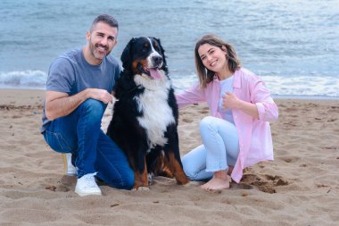 Tatil sırasında kumsalda büyük bir köpekle gülümseyen beyaz bir çiftin portresi. Deniz kenarında rahat ve keyifli bir an. Üçü de aydınlık bir günde kameraya bakıyor.. 