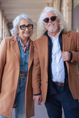 Güneş gözlüğü ve şık ceketler takan gülümseyen yaşlı bir çiftin dikey portresi. Şehir ortamında zarafet, mutluluk ve bağlantı göstererek güvenle dışarı çıkarlar.. 