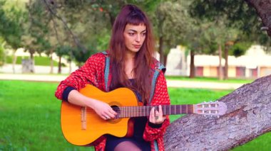 Uzun saçlı genç bir kadın güneşli bir parkta akustik gitar çalıyor, bir ağacın altında huzur içinde oturuyor. Doğal ortam, rahat atmosfer, müzik ve boş zaman atmosferi.