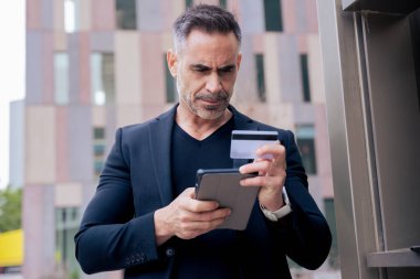 Kafkasyalı yetişkin bir adam tablet kullanıyor ve elinde online ödeme yapmak için bir kredi kartı tutuyor. Modern finans, alışveriş ve dijital yaşam tarzını temsil eden kentsel bir ortamda odaklanmış ifade 