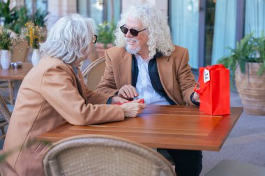 Gözlüklü ve kahverengi paltolu yaşlı bir çift açık hava kafesinde kahvenin tadını çıkarıyor. Sıcak bir anı paylaşıyorlar, gün içinde huzurlu ve rahat bir atmosferde gülümsüyorlar. Dikey