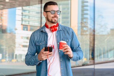 Şehir ortamında akıllı telefonunu kullanırken kırmızı kulaklıklı genç adam kahvenin tadını çıkarıyor. O okurken veya mesajlaşırken gülümsüyor, modern cam binaların önünde sıradan bir anda duruyor.. 