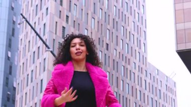 Pembe palto giyen kıvırcık saçlı Afrikalı Amerikalı dansçı şehir ortamında hareketlerini ve enerjilerini ifade ediyor. Modern moda, modern dans ve sanatsal performans.