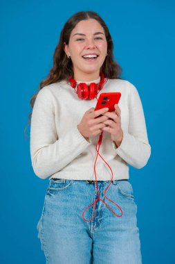 Boynuna kırmızı kulaklık takan, akıllı telefonuna bakıp gülümseyen mutlu beyaz kadın. Mavi arka plan, neşeli ve nişanlı ifadesini vurguluyor. Dikey.