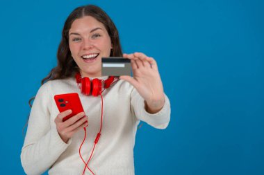 Beyaz kazaklı ve kırmızı kulaklıklı mutlu genç bir kadın kameraya bir kredi kartı gösterirken gülümsüyor ve elinde kırmızı bir akıllı telefon tutuyor, mavi arka planda, online alışverişi sembolize ediyor. Boşluğu kopyala