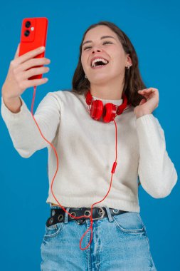 Kırmızı akıllı telefon ve kulaklıkla selfie çeken mutlu genç bir kadının dikey görüntüsü. Mavi bir arkaplan önünde, gündelik ve modern bir dijital anın tadını çıkararak güvenle gülümsüyor. Dikey