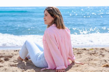 Kumsalda oturmuş, arkasında okyanus varken kameraya gülümseyen mutlu genç bir kadının portresi. Rahat bir ortam, günlük kıyafetler, doğal ışık ve sakin bir ortam.. 