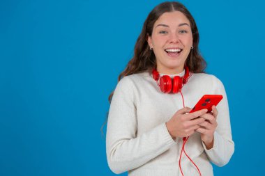 Boynuna kırmızı kulaklık takan, akıllı telefonuna bakıp gülümseyen mutlu beyaz bir kadın. Mavi arka plan onun neşeli ve meşgul ifadesini vurguluyor. Kopya alanı.