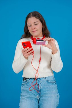 Beyaz kazaklı ve kırmızı kulaklıklı mutlu genç bir kadın kameraya bir kredi kartı gösterirken gülümsüyor ve elinde kırmızı bir akıllı telefon tutuyor, mavi arka planda, online alışverişi sembolize ediyor. Dikey