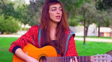 Uzun saçlı genç bir kadın güneşli bir parkta akustik gitar çalıyor, bir ağacın altında huzur içinde oturuyor. Doğal ortam, rahat atmosfer, müzik ve boş zaman atmosferi.