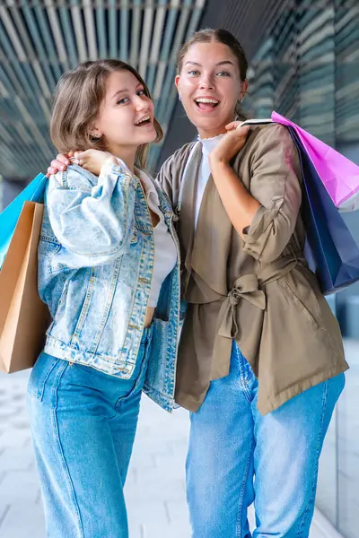 Renkli alışveriş torbaları taşıyan ve alışveriş merkezinde başarılı bir günün ardından gülümseyen iki beyaz kadın. Modern bir binanın dışında duruyorlar, birlikte eğleniyorlar stil ve neşe içinde. Dikey