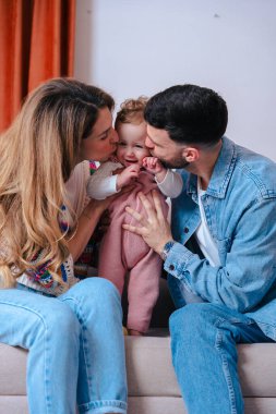 Dikey Portre Mutlu Genç Ebeveynler bebeklerini kanepeye oturtup kameraya gülümsüyorlar. Rahat bir ev ortamında sevgi, sıcaklık ve birliktelikle dolu sıcak aile anları.. 