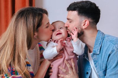 Mutlu ebeveynler evdeki rahat bir kanepede otururken bebeklerini iki yanağından öpüyor. Sahne sıcaklığı, şefkati ve aile bağlarını kapalı bir ortamda yakalıyor.. 