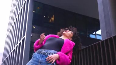 Pembe palto giyen kıvırcık saçlı Afrikalı Amerikalı dansçı şehir ortamında hareketlerini ve enerjilerini ifade ediyor. Modern moda, modern dans ve sanatsal performans.