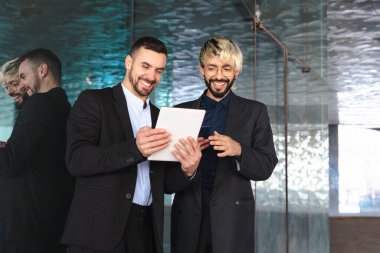 Resmi takım elbiseli iki genç işadamı, bir tablete bakıp gülümserken aynı zamanda şık, modern bir ofiste yansıtıcı yüzeylere ve profesyonel bir atmosfere sahipler.. 