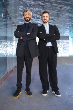 Dikey portre. İki kendine güvenen profesyonel takım elbiseli çapraz kollu adam modern bir ofisin içinde kameraya gülümsüyor. Şirket başarısı, takım çalışması ve iş ortamında liderlik.