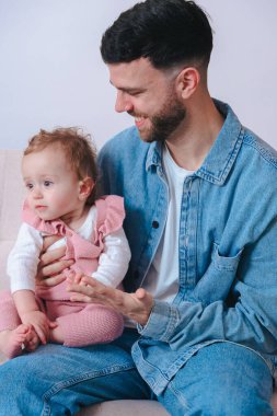 Kot gömlekli genç baba koltuğunda otururken bebeğini nazikçe tutuyor. Bebek, evde sakin ve samimi bir ebeveynlik anını yakalamak için tuhaf bir şekilde uzağa bakıyor.. 