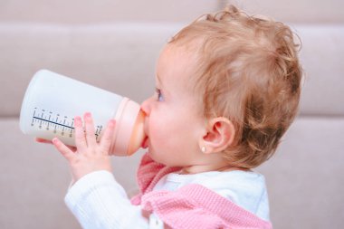 Evde otururken süt şişesini tutan ve içen bir kızın yakın plan görüntüsü. Hassas çocukluk anı, ebeveynlik, beslenme, büyüme ve erken kalkınma temaları için ideal. 