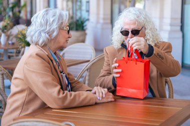 Gözlüklü ve kahverengi paltolu yaşlı bir çift açık hava kafesinde kahvenin tadını çıkarıyor. Sıcak bir anı paylaşıyorlar, gün içinde huzurlu ve rahat bir atmosferde gülümsüyorlar. Dikey