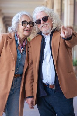 Dışarıda mutlu son sınıf çifti. Kıvırcık beyaz saçlı ve güneş gözlüklü adam kamerayı işaret ediyor, gülümsüyor, gözlüklü kadın ise yanında duruyor, hem neşeli hem de kendinden emin bir şekilde..