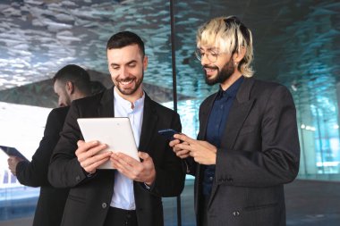 Resmi takım elbiseli iki genç işadamı, bir tablete bakıp gülümserken aynı zamanda şık, modern bir ofiste yansıtıcı yüzeylere ve profesyonel bir atmosfere sahipler.. 