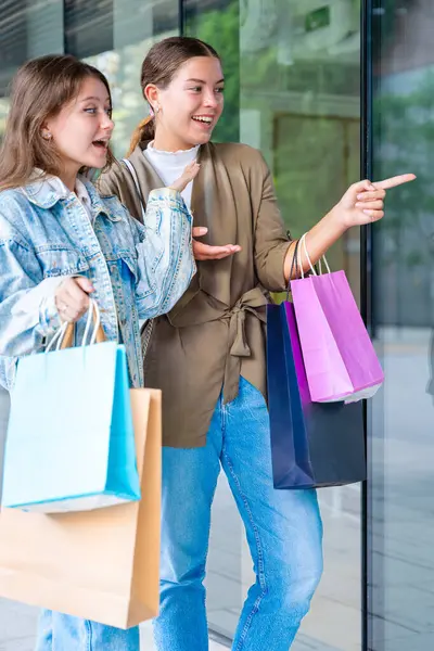Alışveriş torbaları tutan ve vitrinlere bakan iki mutlu beyaz kadın. Şehirde alışveriş ve arkadaşlığın olduğu bir günde samimi ve neşeli bir şehir anı. Dikey
