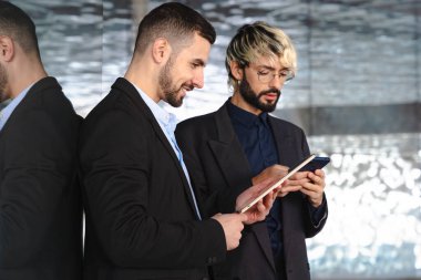 Takım elbiseli iki genç işadamı, modern, şık bir ofis alanında yansıtıcı cam duvarlar ve profesyonel, teknoloji odaklı bir atmosferle işbirliği yaparken dijital tabletlere odaklandılar.. 