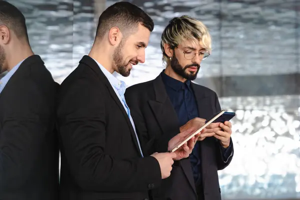 Takım elbiseli iki genç işadamı, modern, şık bir ofis alanında yansıtıcı cam duvarlar ve profesyonel, teknoloji odaklı bir atmosferle işbirliği yaparken dijital tabletlere odaklandılar.. 