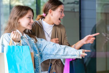 Alışveriş torbaları tutan ve vitrinlere bakan iki mutlu beyaz kadın. Şehirde alışveriş ve arkadaşlığın olduğu bir günde samimi ve neşeli bir şehir anı. 